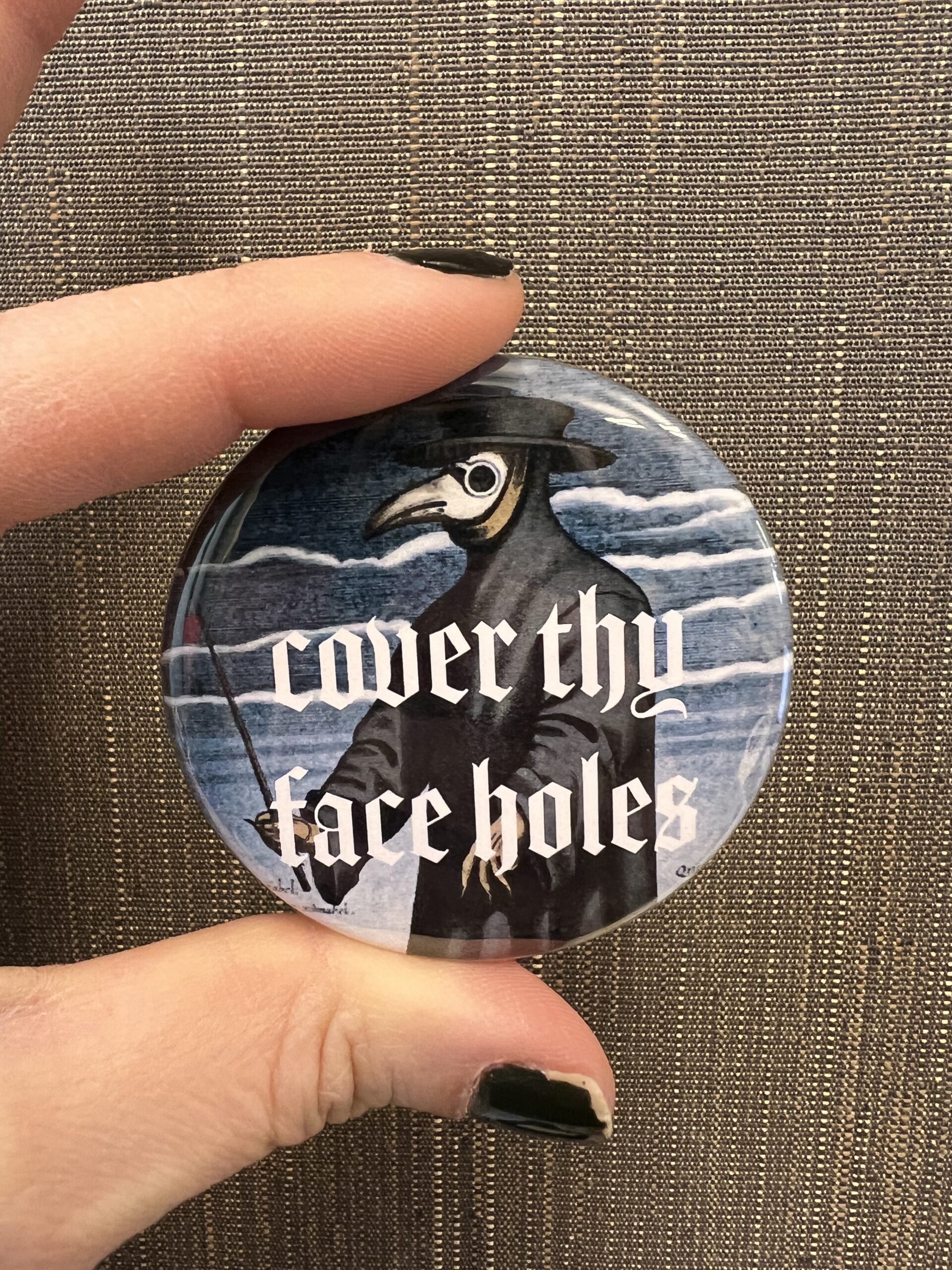 Photo of two fingers with black nail polish holding a round pin. The pin shows a figure in a plague mask, with the words 