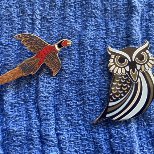 A brown, black, red and white pheasant pin and a brown and white owl pin